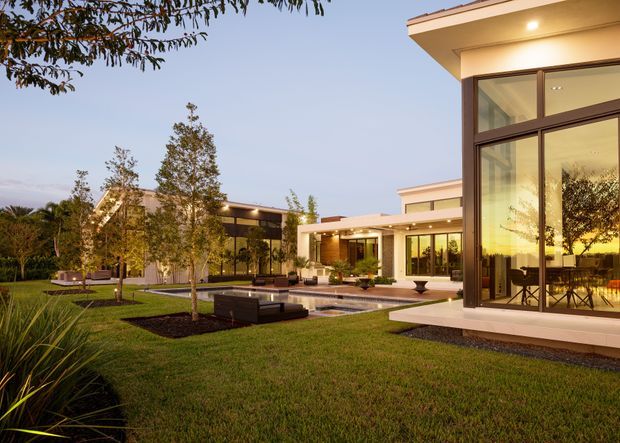 A spacious contemporary residential courtyard viewed from the landscaped garden at dusk. The home features crisp flat rooflines, pale rendered walls, extensive dark-framed floor-to-ceiling glazing, and multiple connected pavilion-like volumes around a paved outdoor entertaining area. Lawn, ornamental planting, built-in seating, and warm interior lighting create a refined indoor-outdoor setting.