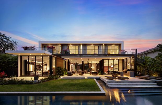 Twilight exterior view of a large two-storey contemporary residence with a flat roof, extensive floor-to-ceiling glazing, warm interior lighting, open terraces and balconies, landscaped lawn, broad steps, and a reflective swimming pool in the foreground.
