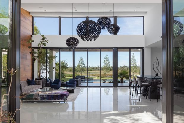 A spacious, double-height contemporary open-plan interior with a living area, dining zone and kitchen visible together. Full-height glazing opens to a landscaped outlook and reflects abundant daylight across the polished pale floor. The room features a warm timber-clad accent wall, dark-framed glazing, sculptural black woven pendant lights, neutral upholstered seating, indoor plants and a dining table with bar-height stools.