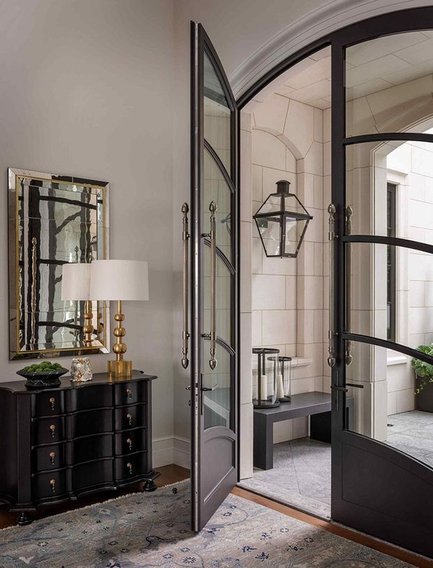 A refined entryway with a dark traditional dresser, brass table lamp, decorative mirror, patterned rug, and ornate black-framed glazed double doors opening to a bright stone-clad exterior vestibule. The doors feature curved transoms and decorative metal hardware, creating a formal, elegant transition between interior and exterior.
