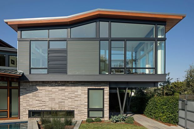 Contemporary two-storey residential facade with extensive floor-to-ceiling glazing, dark horizontal cladding, pale brickwork, and a projecting upper level supported by angled structural posts. The low-pitched roof has broad eaves with a warm timber-toned soffit or fascia. A landscaped entry path and a glimpse of a pool appear in the foreground.