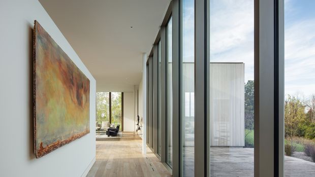 A bright, refined contemporary interior hallway or circulation space with full-height black-framed glazing along one side, pale timber flooring, white walls, and a large abstract artwork. The corridor leads toward a furnished living area and opens visually to an exterior deck and landscaped setting through the glazing.