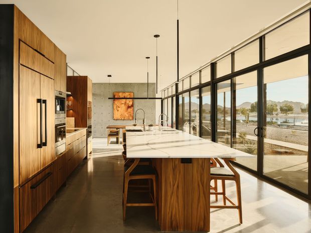 A spacious contemporary kitchen with a long central island, integrated appliances, and extensive floor-to-ceiling black-framed glazing. Warm timber cabinetry contrasts with a white stone island and polished concrete surfaces, while pendant lights and exposed concrete add a refined modern character. The glazing opens to a bright outdoor landscape with hills and water visible beyond.