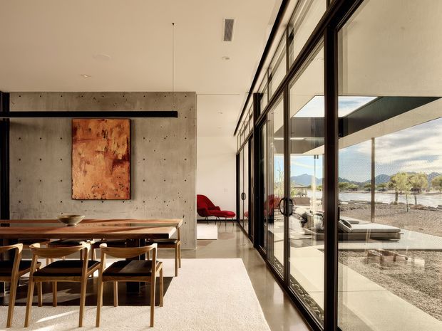 Contemporary open-plan dining and living space with a long timber dining table and wooden chairs positioned beside full-height black-framed glazing. A raw concrete feature wall with an abstract artwork contrasts with the warm timber furniture and light polished floor. The glazing opens visually to a covered outdoor terrace and a mountainous landscape beyond.