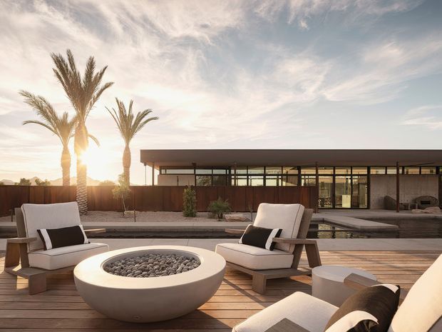 A refined outdoor lounge area on a timber deck, featuring a large round fire bowl filled with stones, upholstered lounge chairs, and a low outdoor table. Beyond the deck is a long, low modern residence with extensive glazing, dark horizontal rooflines, timber screening, a reflecting pool, gravel landscaping, and tall palm trees in warm late-afternoon light.