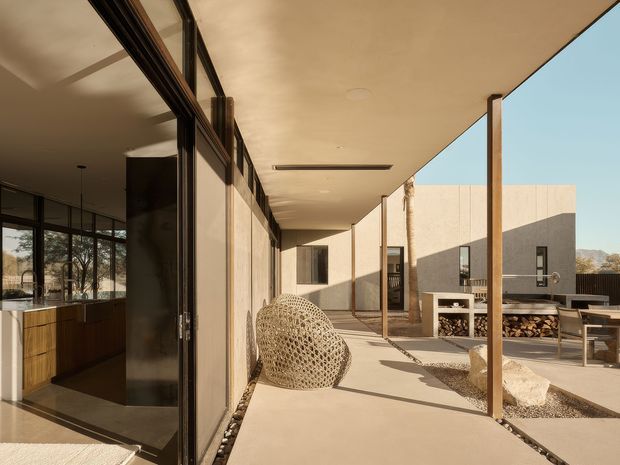 A covered outdoor terrace or courtyard beside a modern home, viewed along floor-to-ceiling glazed walls. The space has a smooth concrete terrace floor, slender timber posts, warm neutral plastered walls, a built-in outdoor fireplace with stacked firewood, a dining table and chairs, and a woven lounge chair. The adjoining interior kitchen is visible through the glazing, while distant landscape appears beyond the courtyard.