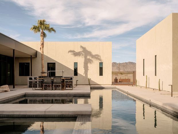 A minimalist modern courtyard centered around a long rectangular swimming pool, with pale cream rendered walls, narrow vertical windows, a shaded outdoor dining terrace, and a tall palm casting a shadow across the facade. Mountain terrain is visible beyond the property, reinforcing the warm desert-like setting. The architecture has crisp geometric volumes and restrained detailing, with reflective water and pale paved surfaces creating a calm, sunlit atmosphere.