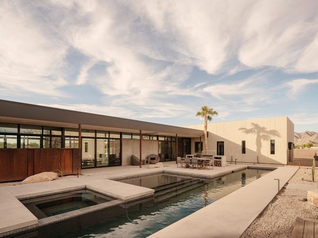 A low-slung modern residence surrounding a long rectangular swimming pool and attached spa. The building has broad floor-to-ceiling glazing, a flat roof, dark metal-framed windows, pale rendered walls, and a covered outdoor dining and lounge area. Desert-style gravel landscaping, a palm tree, and distant rocky hills are visible beyond the pool.