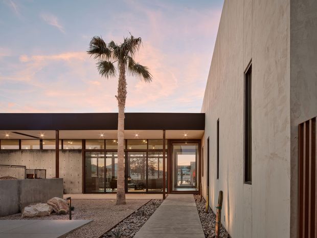 A contemporary courtyard-style exterior at sunset, with a low horizontal pavilion featuring extensive floor-to-ceiling glazing and dark metal framing. A tall rendered wall lines the right side, while a concrete pathway passes through gravel and stone landscaping toward glazed doors. A palm tree and an outdoor seating/fireplace area are visible in the courtyard.