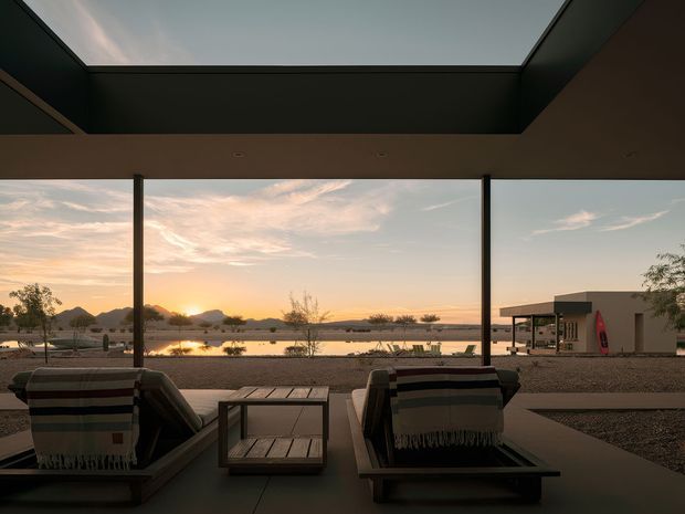 A covered outdoor terrace frames a sunset view over a calm lake, with two low lounge chairs draped in striped throws and a small square coffee table in the foreground. Slim dark posts support a broad roof with a large rectangular skylight or opening. Across the water is a sparse, low-lying landscape and a minimalist pavilion-like building, creating a serene, modern waterside setting.
