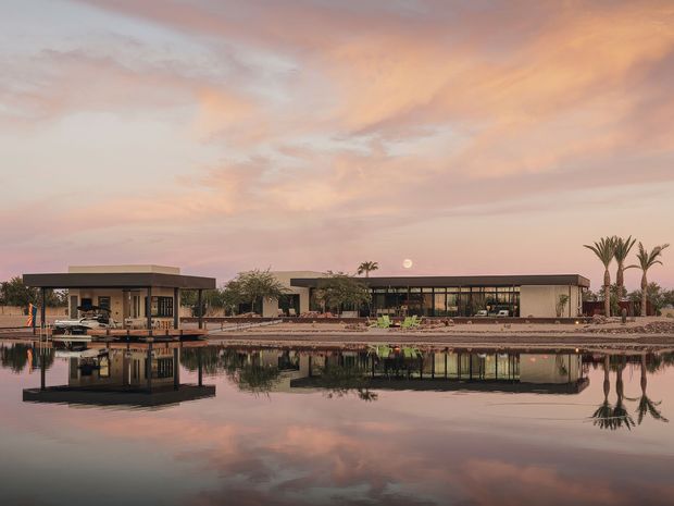 Wide exterior view of a low, contemporary single-storey building arranged around a landscaped waterfront or reflective pond. The structure features broad flat roof planes, extensive floor-to-ceiling glazing, dark slender framing, and pale rendered walls. A covered terrace or pavilion projects over the water at left, while palms and carefully landscaped planting surround the building. The calm sunset sky and strong reflections create a serene, luxurious atmosphere.