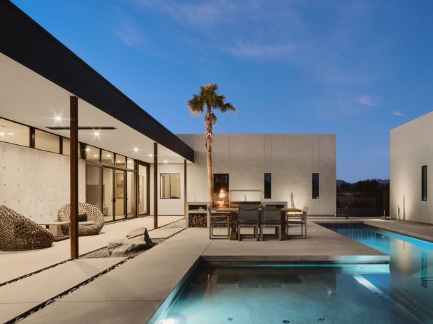 A modern single-level courtyard-style home viewed from an outdoor terrace at dusk. The scene features crisp white rendered walls, extensive floor-to-ceiling glazing, a broad dark roof or fascia edge, a concrete-paved terrace, outdoor dining furniture, a palm tree, and a rectangular swimming pool with an illuminated shallow section. The architecture has strong horizontal lines and minimalist detailing.