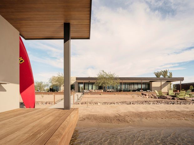 A low-slung modern building viewed from a covered timber deck, with broad glazing, a long projecting flat roof and slender dark structural columns. The landscaped setting is arid and desert-like, with gravel beds, sparse planting and sandy ground. A bright red surfboard-like object stands at the left edge, while the composition emphasises the building's horizontal lines and connection to the outdoor space.
