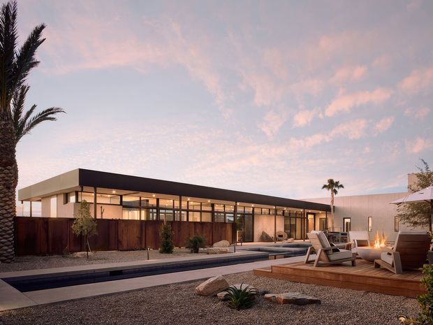 Single-storey contemporary residence photographed at dusk, with a long low flat-roofed form, extensive floor-to-ceiling glazing, dark metal framing, and pale rendered walls. A narrow linear swimming pool runs alongside the building, while a timber deck with outdoor loungers and a small fire feature occupies the foreground. Gravel landscaping, palms, and sparse desert-style planting reinforce the warm modern resort-like setting.