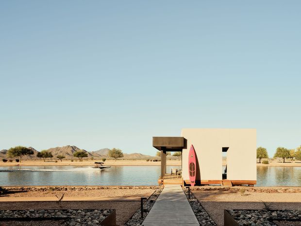 A minimalist waterfront pavilion or small modern structure beside a calm body of water in an arid desert landscape. A straight timber walkway crosses a rocky landscaped foreground toward the building, with a shaded timber entry canopy and a bright pink surfboard or paddleboard leaning against the facade. Low mountains, sparse trees, and sandy terrain form the background under a clear blue sky.