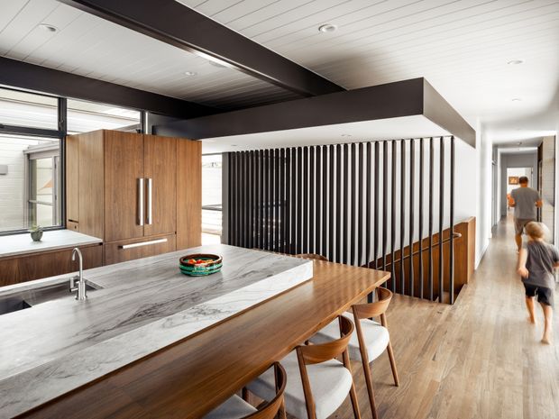 Open-plan contemporary kitchen and dining area with a large white-and-grey stone island, timber cabinetry, a timber dining table with stools, and a dark vertical slatted divider. The space has light timber flooring, a white linear-panel ceiling with exposed dark structural beams, extensive glazing, and a hallway extending to the right.