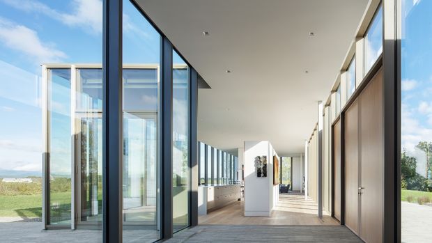 A bright, expansive contemporary interior corridor or open-plan living space with floor-to-ceiling glazing on both sides, slim dark window frames, pale timber flooring, white walls and ceiling, and integrated light-wood cabinetry. The space opens toward a landscaped exterior and distant coastal-looking landscape, with strong natural light and a clean minimalist composition.