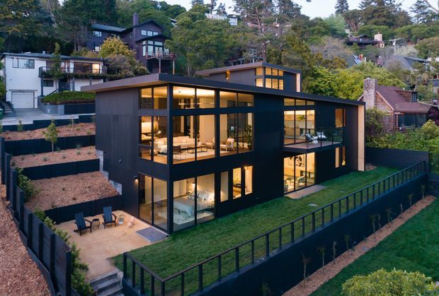 A modern multi-level residential house on a steep, landscaped suburban site. The dark-clad building has extensive floor-to-ceiling glazing, projecting flat roof planes, and warm illuminated interiors visible from outside. Terraced retaining walls, planted roofs or lawns, external stairs, decks, and black metal balustrades articulate the sloping garden.