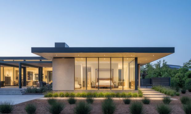 Contemporary single-storey house exterior at dusk, featuring a strong flat projecting roof, extensive floor-to-ceiling glazing, warm illuminated interiors, and a landscaped front garden with ornamental grasses and paved paths. The image primarily presents the building facade and its indoor-outdoor connection.