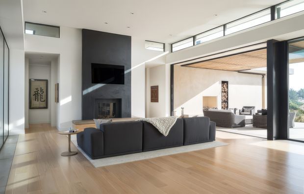 Bright contemporary living room with pale timber flooring, a large dark charcoal sectional sofa, and a tall dark feature wall containing a recessed television and fireplace. Full-height sliding glass doors open to a covered outdoor lounge with a timber-lined ceiling and built-in fireplace, while clerestory glazing brings in abundant daylight.