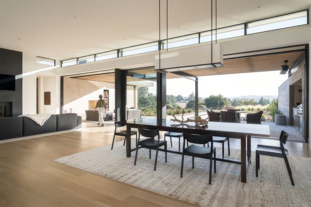 A bright, open-plan contemporary living and dining space with extensive floor-to-ceiling glazing and clerestory windows. A long timber dining table with black chairs sits on a large pale rug, while a dark sectional sofa and media wall occupy the left side. The room opens directly to an outdoor terrace and landscaped view, with a striking linear pendant light and a timber-lined ceiling section adding warmth.