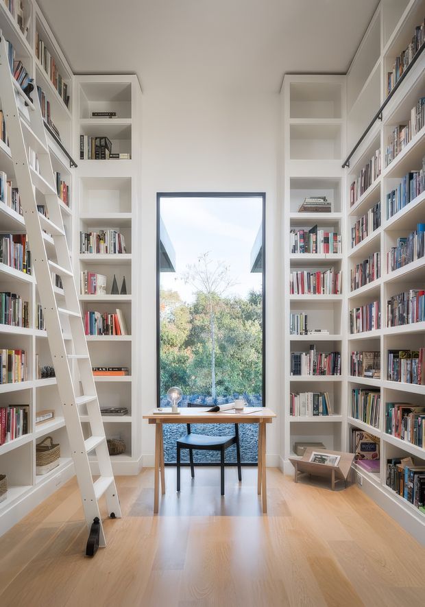 A bright, narrow home library lined on both sides with floor-to-ceiling white built-in bookshelves filled with books. A rolling ladder leans against the left shelving, while a large black-framed window overlooks greenery at the far end. A simple timber desk and black chair sit in front of the window, with pale timber flooring and predominantly white walls creating an airy, organized space.