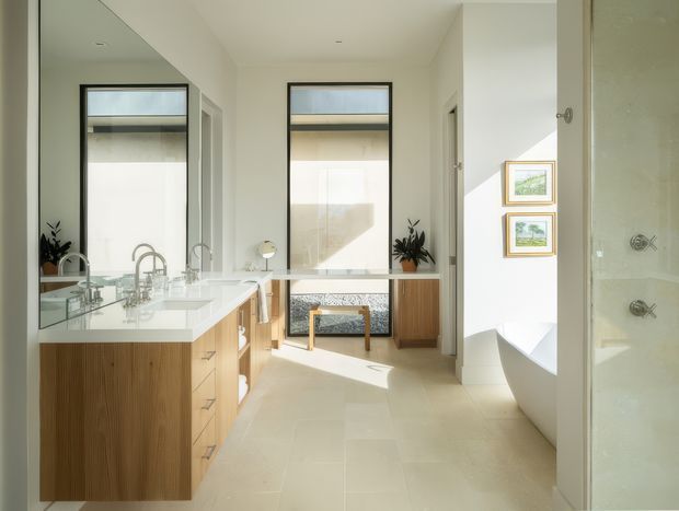 Bright, spacious contemporary bathroom with a long double vanity, mirrored wall, white integrated basins, and warm vertical-grain timber cabinetry. Large windows bring in abundant daylight, while a freestanding white bathtub is partially visible on the right beside a glass-enclosed shower. Pale large-format floor tiles, white walls, minimal fittings, and framed artwork create a clean, refined atmosphere.