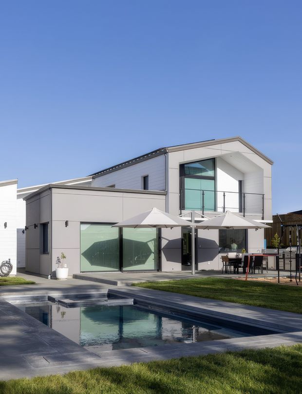 A bright contemporary two-storey home with a crisp white and light-grey facade, flat and gently pitched roof forms, large glazed openings, and a glass-balustraded upper balcony. The landscaped backyard includes a rectangular swimming pool with dark paving, outdoor dining, and multiple light-coloured umbrellas.