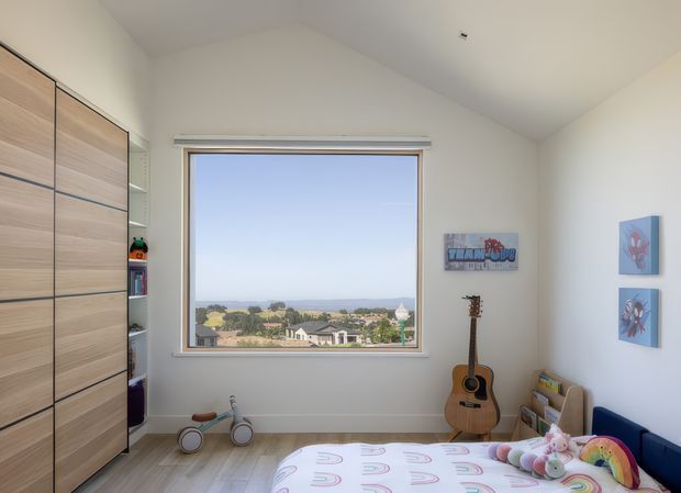 Bright children's bedroom with a large picture window overlooking a suburban landscape. The room contains full-height light timber built-in wardrobes and shelving, a bed with playful rainbow-patterned bedding and soft toys, a small tricycle, a guitar, children's artwork, and book storage. The space has white walls, a simple white ceiling, and abundant natural light.