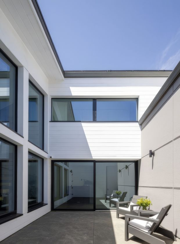 Bright contemporary courtyard formed by a white, multi-level house. The enclosed outdoor terrace has dark concrete paving, large black-framed glazed doors and windows, white horizontal cladding, and a contrasting rendered wall with slim wall lights. Two modern outdoor lounge chairs and a small table occupy the sunlit foreground, while strong angular shadows emphasize the geometric architecture.