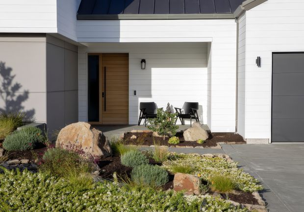 Contemporary residential front entrance with white horizontal weatherboard cladding, a dark metal roof edge, timber pivot-style front door, and an attached garage. Two outdoor chairs sit in a recessed entry porch, which opens onto a landscaped drought-tolerant garden with large decorative rocks, gravel, and low planting.