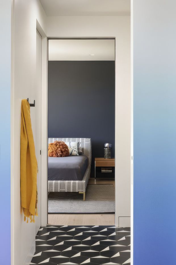 View through a narrow interior hallway into a compact bedroom. The hallway has a bold black-and-white geometric floor, white walls, and a vivid blue door or wall panel on the right. A mustard-yellow textile hangs from a wall hook. The bedroom beyond features a dark charcoal feature wall, a low upholstered bed with a patterned frame, a small wooden bedside table, a metallic lamp, and a neutral rug. The composition is clean and contemporary with strong graphic contrast.