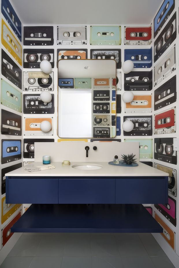 A compact, highly graphic bathroom featuring walls covered in colourful cassette-tape imagery, a large mirrored medicine cabinet, spherical wall lights, and a floating deep-blue vanity with a white benchtop and integrated basin. The room has a playful retro-inspired aesthetic, dark blue flooring, and a grey tiled floor area.