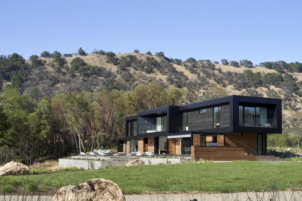 A contemporary two-storey house set against a dry, wooded hillside. The building has a bold dark rectangular form with extensive glazing, projecting upper-level volumes, warm timber-clad sections, and a broad lawn and terrace in front.