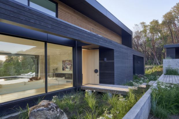 Modern single-storey residential exterior with strong horizontal massing, dark horizontal cladding, warm timber wall panels, expansive floor-to-ceiling glazing, and a recessed timber entry door. The building is set within a landscaped garden with native grasses, rocks, concrete retaining edges, and a timber walkway. Interior living spaces are visible through the glazing.