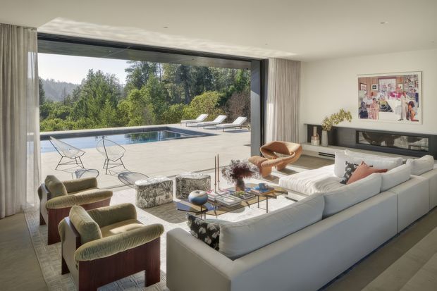 A bright, spacious contemporary living room with floor-to-ceiling retractable glazing opening onto a large paved pool terrace and landscaped garden. The room features a large white sectional sofa, sculptural upholstered lounge chairs, a glass coffee table, a linear fireplace or dark built-in feature wall, neutral walls, sheer curtains, and framed artwork. The open indoor-outdoor connection dominates the composition.