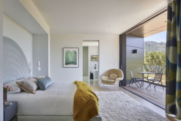 Bright, refined bedroom with a sculptural curved upholstered feature headboard, white bedding, soft neutral furnishings, and a mustard throw. The room opens through large sliding glass doors to a timber deck with outdoor seating and views of a wooded hillside. A compact adjoining bathroom or vanity area is visible through the central opening, while patterned curtains frame the glazing.