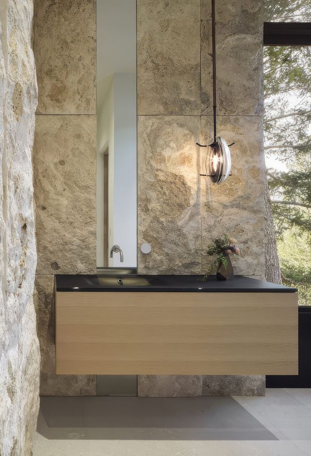 A refined contemporary bathroom vanity viewed straight on. The floating timber vanity has a dark stone benchtop with an integrated basin and wall-mounted tapware. Behind it, large-format textured natural stone wall panels create a strong feature surface, complemented by a slim vertical mirror, sculptural wall light, and a full-height window revealing trees outside. The palette is warm and earthy with minimalist detailing.