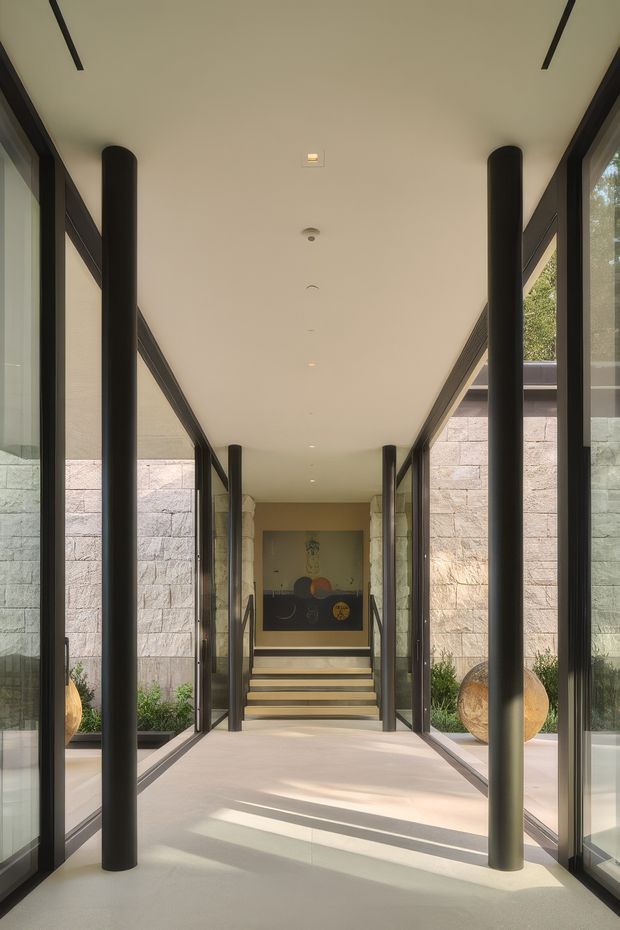 A long, symmetrical contemporary entry corridor framed by full-height dark glazing and slender black cylindrical columns. Pale stone block walls, a smooth light floor, and a recessed white ceiling create a calm, gallery-like passage leading to a short stair and artwork at the far end. Strong natural light enters from the glazed sides, with planted areas and sculptural objects visible beyond the glass.