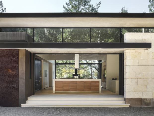 Front exterior view of a modern, single-storey residence with a broad flat roof, extensive black-framed glazing, and a recessed open kitchen visible behind a series of entry steps. The facade combines pale stone masonry, dark metal framing, concrete-like horizontal bands, and a textured dark feature wall, with mature trees visible through the glazing.