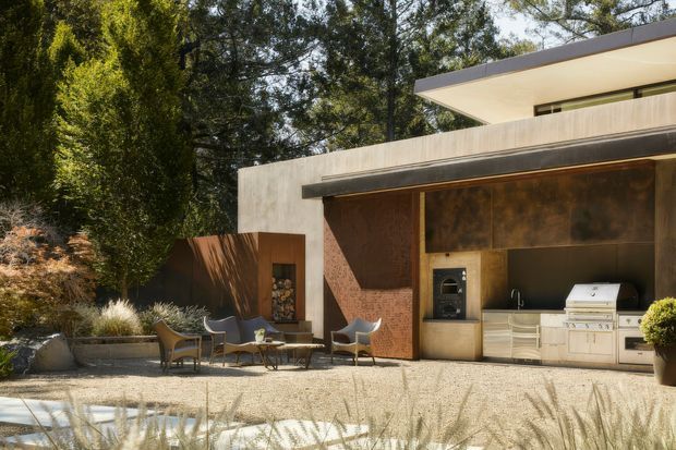 A sophisticated outdoor entertaining area beside a modern home, featuring a built-in barbecue kitchen beneath a deep roof overhang. The space has warm textured masonry and timber-toned surfaces, pale built-in cabinetry, a stone or concrete-paved terrace, and a small seating arrangement surrounded by mature landscaping. The building has strong horizontal lines, a low-pitched roof, and large sheltered openings.