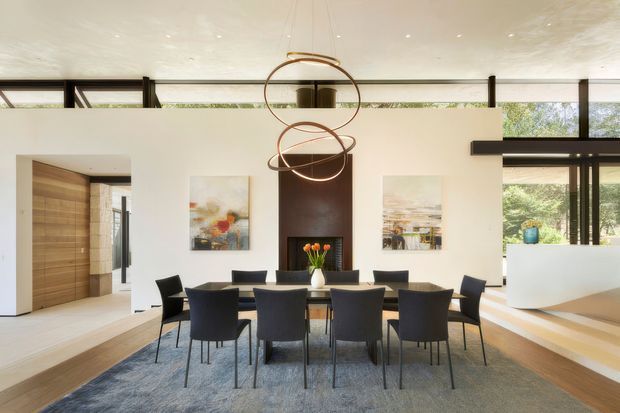 Bright contemporary dining room with a long dark dining table and multiple upholstered chairs, centred on a tall dark feature wall with a fireplace and flanked by abstract artwork. The room has white walls and ceiling, warm timber flooring and door joinery, expansive black-framed glazing, sculptural suspended ring lighting, and a stepped transition toward an adjacent living area.