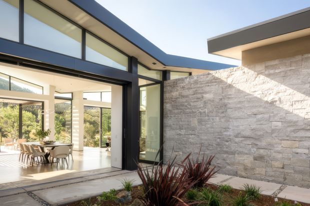 A contemporary flat-roofed home viewed from an outdoor courtyard or entry area. The facade combines expansive black-framed glazing with pale rendered surfaces and a prominent textured light-stone wall. Through the open glazing, a bright dining area with a long table and chairs is visible. Minimal landscaping with sculptural grasses sits in the foreground, and strong sunlight creates a clean, warm architectural presentation.