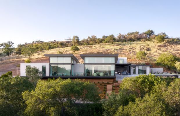 A modern single-storey hillside residence viewed from the surrounding landscape. The home has a low, linear composition with broad floor-to-ceiling glazing, pale rendered walls, dark horizontal roof planes and framing, and a central outdoor terrace or circulation zone. Timber-toned steps descend through dense foreground vegetation, while the dry, gently sloping site rises behind the house.