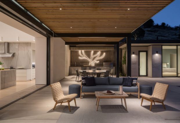A sophisticated open-plan indoor-outdoor living and dining area at dusk. The covered terrace features a timber slatted ceiling, dark structural framing, a deep blue sofa with matching ottomans, woven lounge chairs, and a timber coffee table. Beyond is a dining zone with a sculptural illuminated wall feature, while large retractable glazed openings connect directly to a refined kitchen with pale cabinetry and a stone-look splashback. The space has warm integrated lighting and a strong connection to the courtyard and night landscape.