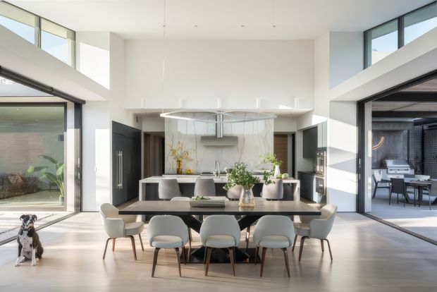 Bright, spacious contemporary open-plan kitchen and dining area with a large island, dark cabinetry, marble-look splashback, and a long dining table surrounded by upholstered chairs. Floor-to-ceiling glazing and high clerestory windows create strong natural light, with an outdoor entertaining area visible through the opening on the right. The room has a high ceiling and a refined neutral palette with dark accents.