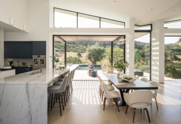 Bright, open-plan contemporary kitchen and dining area with a large marble-look island, dark built-in cabinetry, bar seating, and a long dining table. Floor-to-ceiling glazing and clerestory windows frame a landscaped hillside, while strong sunlight creates linear shadows across the pale floor. The space opens directly to an outdoor terrace and pool area.