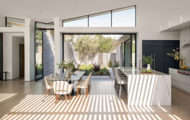 Bright contemporary open-plan kitchen and dining area with a marble-look island, dark integrated cabinetry, a timber dining table with upholstered chairs, and extensive floor-to-ceiling glazing opening toward a planted courtyard. Strong sunlight creates dramatic linear shadows across the pale floor.