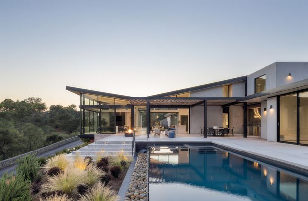 Contemporary multi-wing residence viewed from the landscaped pool terrace at dusk. The home features broad floor-to-ceiling glazing, crisp white rendered walls, dark metal framing, low-slung projecting roof planes, covered outdoor living areas, and a rectangular swimming pool bordered by pale paving and rounded stone landscaping. Warm interior lighting creates a refined indoor-outdoor connection, with trees and planted grasses surrounding the site.