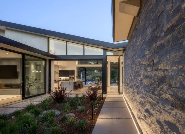 Modern residential courtyard viewed from a paved walkway, with a prominent dark stacked-stone wall on the right and floor-to-ceiling black-framed glazing opening into a bright kitchen and living area. The low, angular roofline and clerestory glazing surround a landscaped courtyard planted with ornamental grasses and burgundy accent plants.
